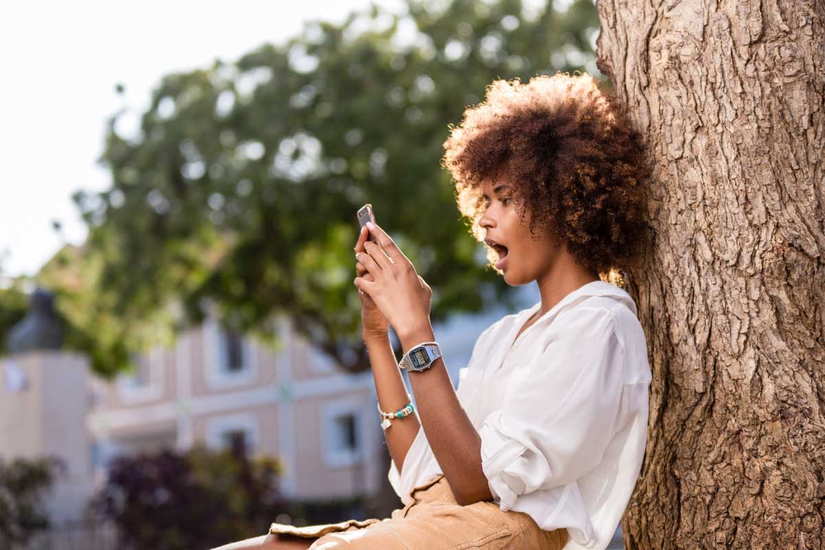 Mujer joven, sentada y recostada en un árbol grueso, mirando sorprendida la pantalla de un teléfono celular.
