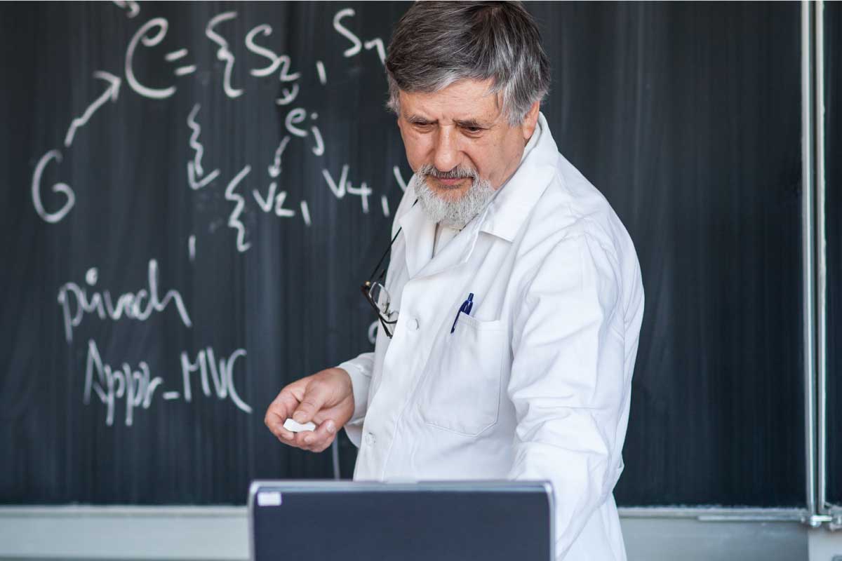 Profesor al frente de un computador, con una tiza en su mando y un tablero con ecuaciones en el fondo