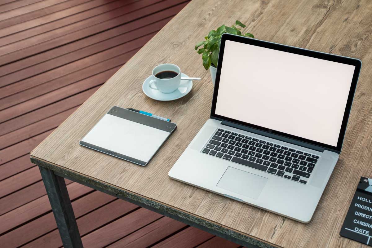 Computador portatil abierto sobre escritorio en el que además hay una tableta, un café y una planta