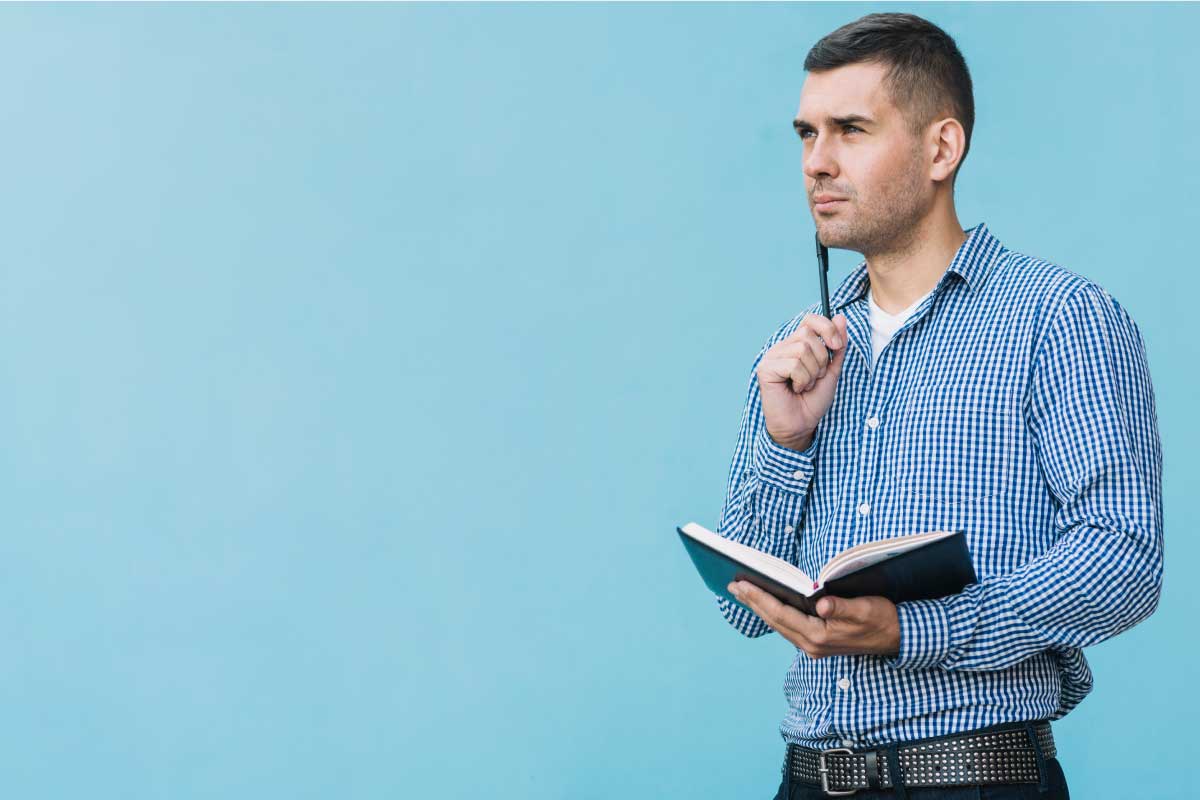 Hombre joven, parado al frente de una pared azul, sosteniendo una libreta y un lápiz sobre la mejilla, pensando.