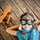 Niño vestido con gorra y gafas de piloto, acostado sobre un piso de madera con sus manos bajo su cabeza y sonriendo.