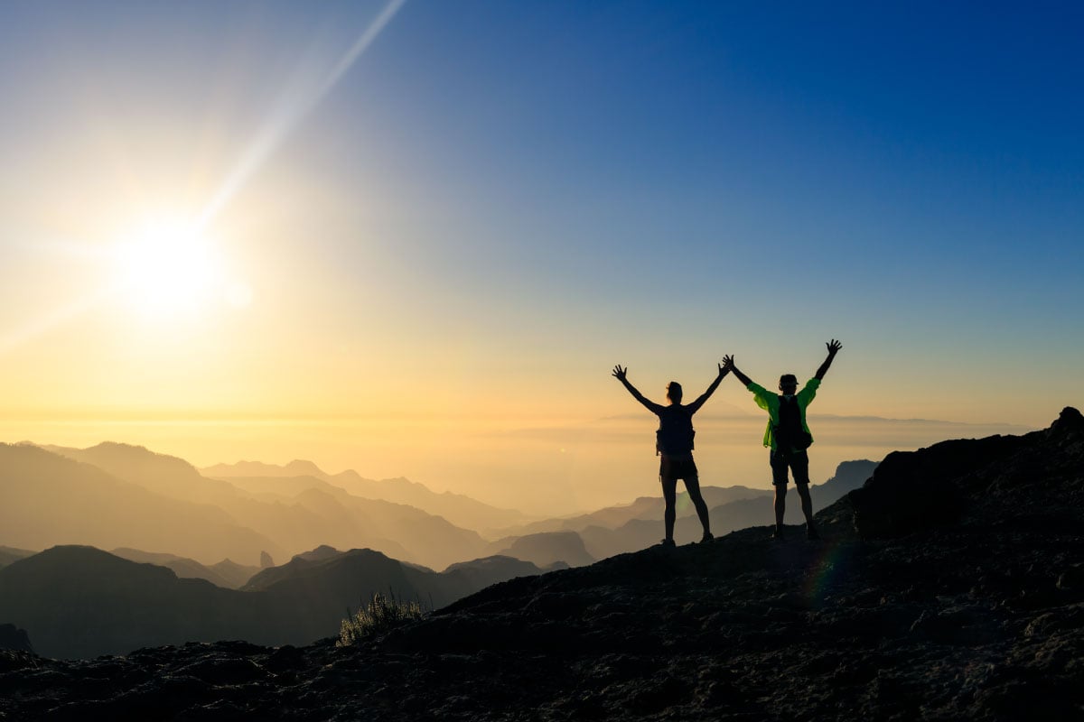 Dos personas en la cima de una montaña con las manos levantadas hacia el cielo.