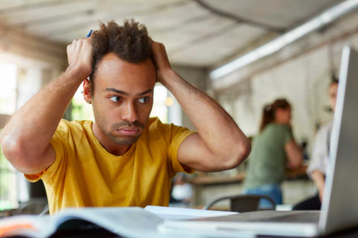 Hombre joven sentado al frente un computador portatil, con sus manos en la cabeza en signo de frustración