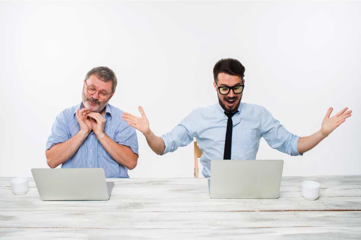 Toma frontal de dos hombres cada uno sentado al frente de un computador portatil, uno con los brazos abiertos y sonriendo.