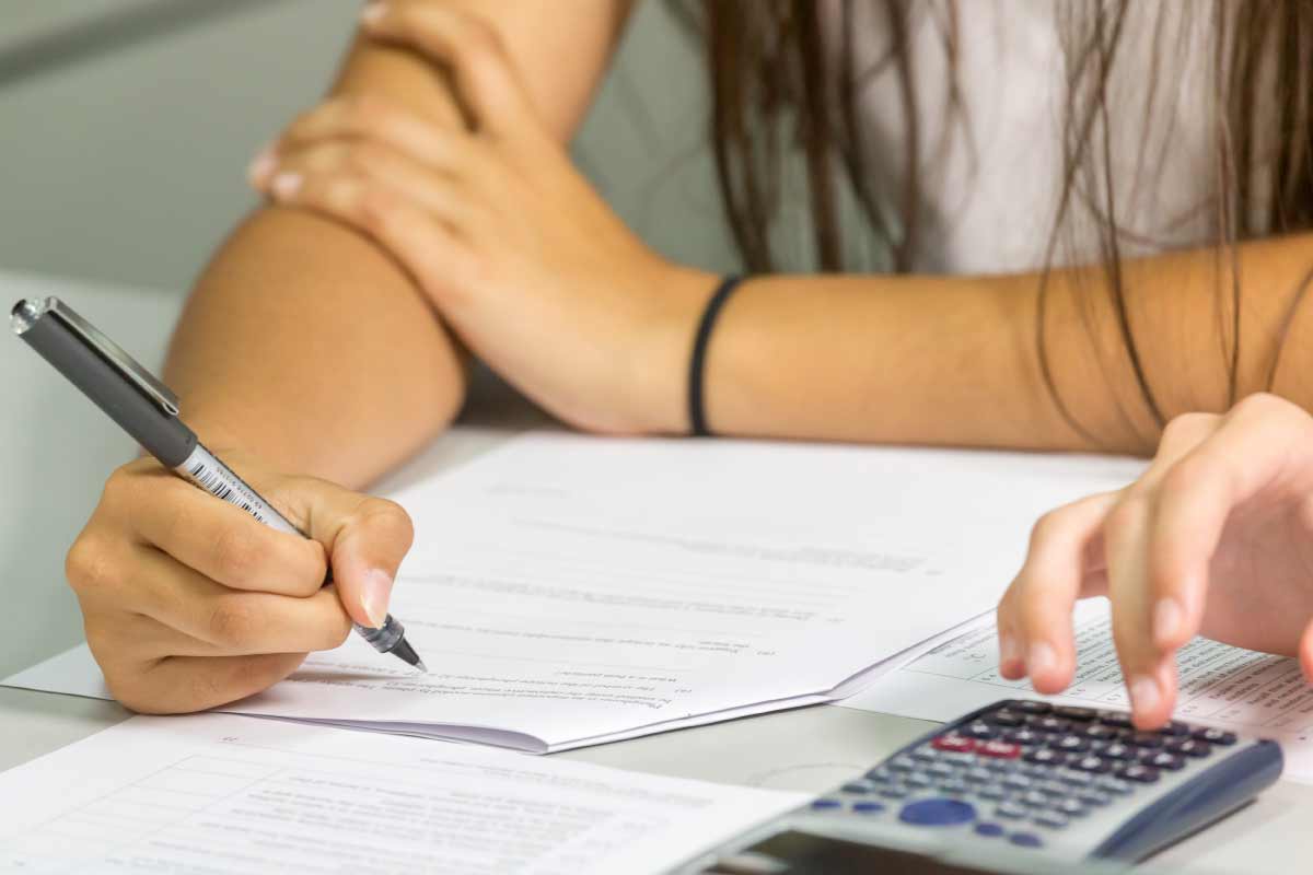 Acercamiento de las manos de dos estudiantes, uno llenando un formulario en papel, el otro usando una calculadora.