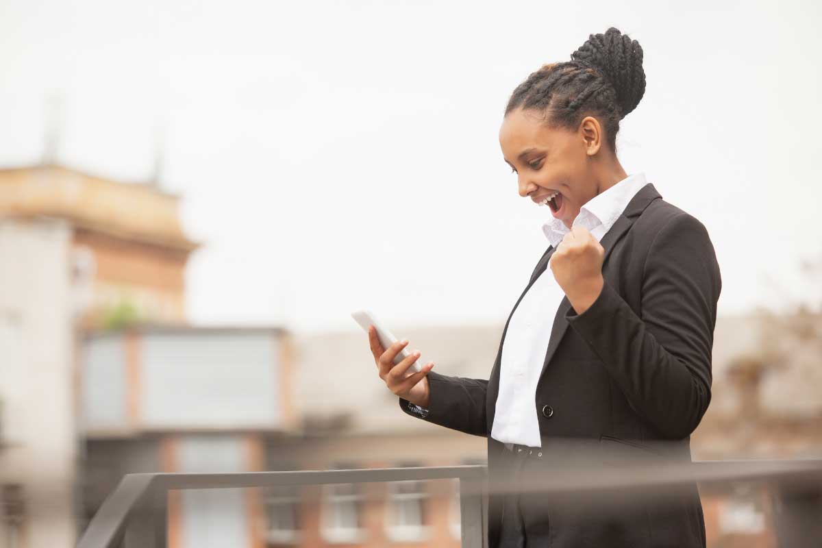 Mujer joven en un balcón, leyendo un teléfono móvil, empuñando su otra mano y con expresiones de victoria en su cara.
