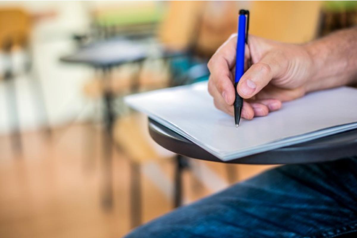 Acercamiento de la mano de un estudiante sentado en un pupitre y escribiendo en un cuaderno.