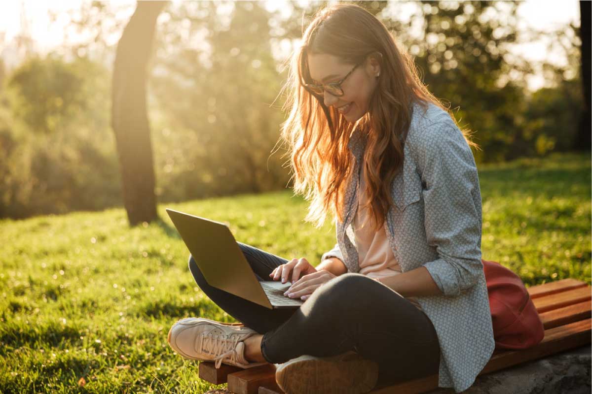 Mujer joven sentada en un parque con los pies cruzados escribiendo en un computador portatil.