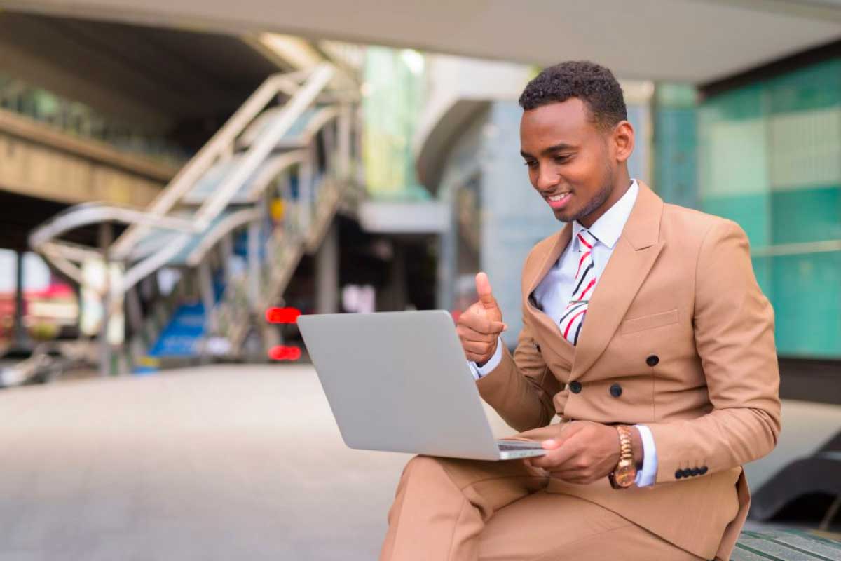 Hombre de negocios sentado en una estación de transporte con un computador portatil sobre sus piernas y levantando el dedo pulgar al frente de la pantalla.