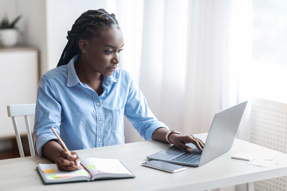Mujer joven sentada al frente de un escritorio, mirando un computador portable y tomando notas en una libreta.