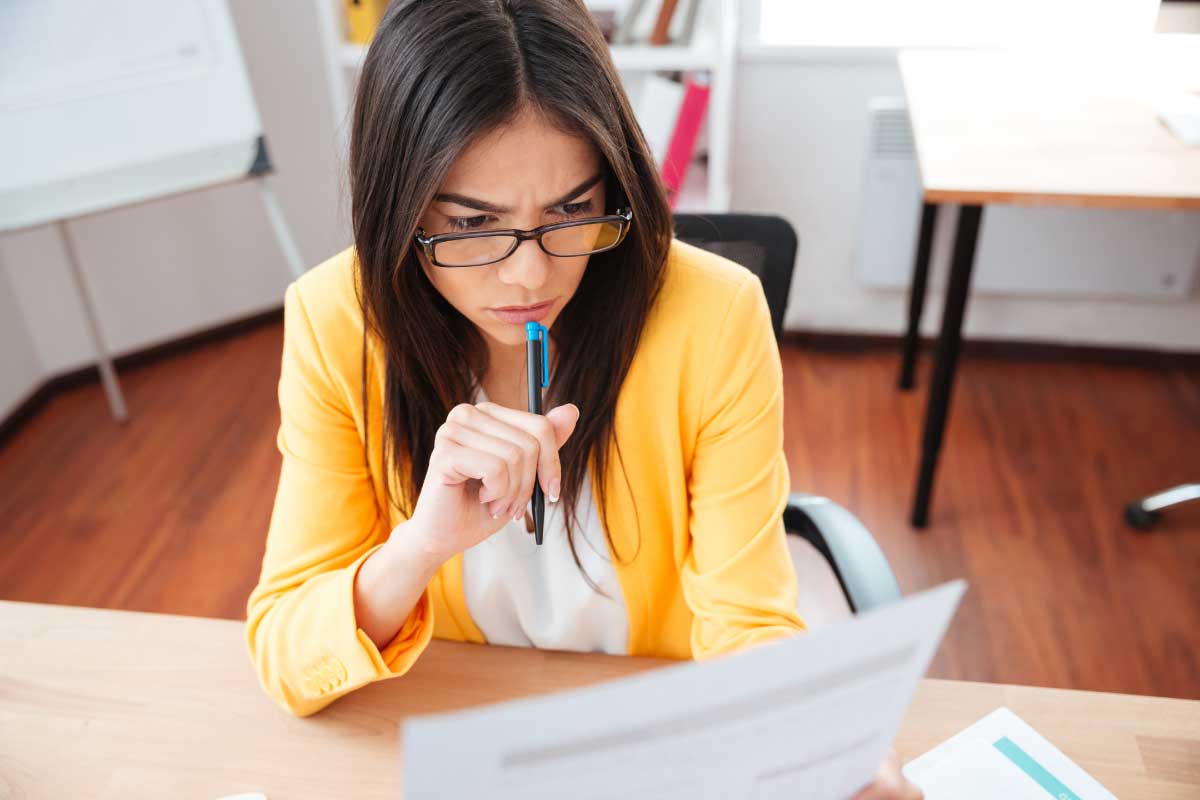 Mujer joven revisando atentamente documentos en papel, sosteniendo un lapicero sobre sus labios.