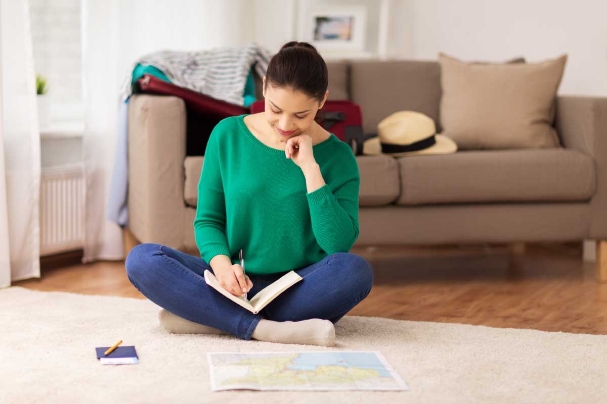 Mujer joven, sentada en el piso de la sala de su casa, tomando nota en una libreta y un mapa en papel a su lado.