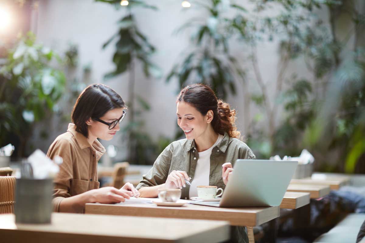 Dos mujeres jóvenes sentadas en una cafetería, con un computador portatil abierto y revisando notas.