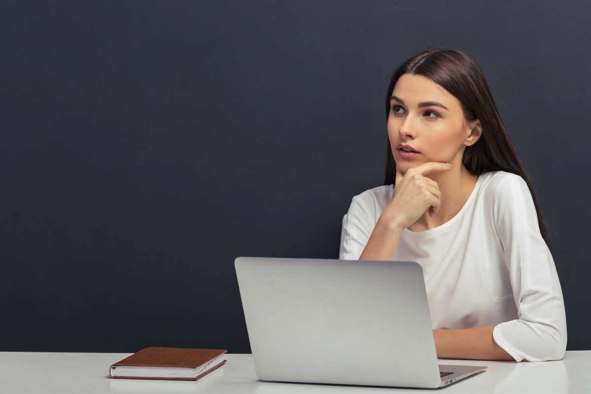 Mujer joven sentada al frente de un escritorio, con un computador portable abierto, su mano sobre su mentón mirando hacia un lado.