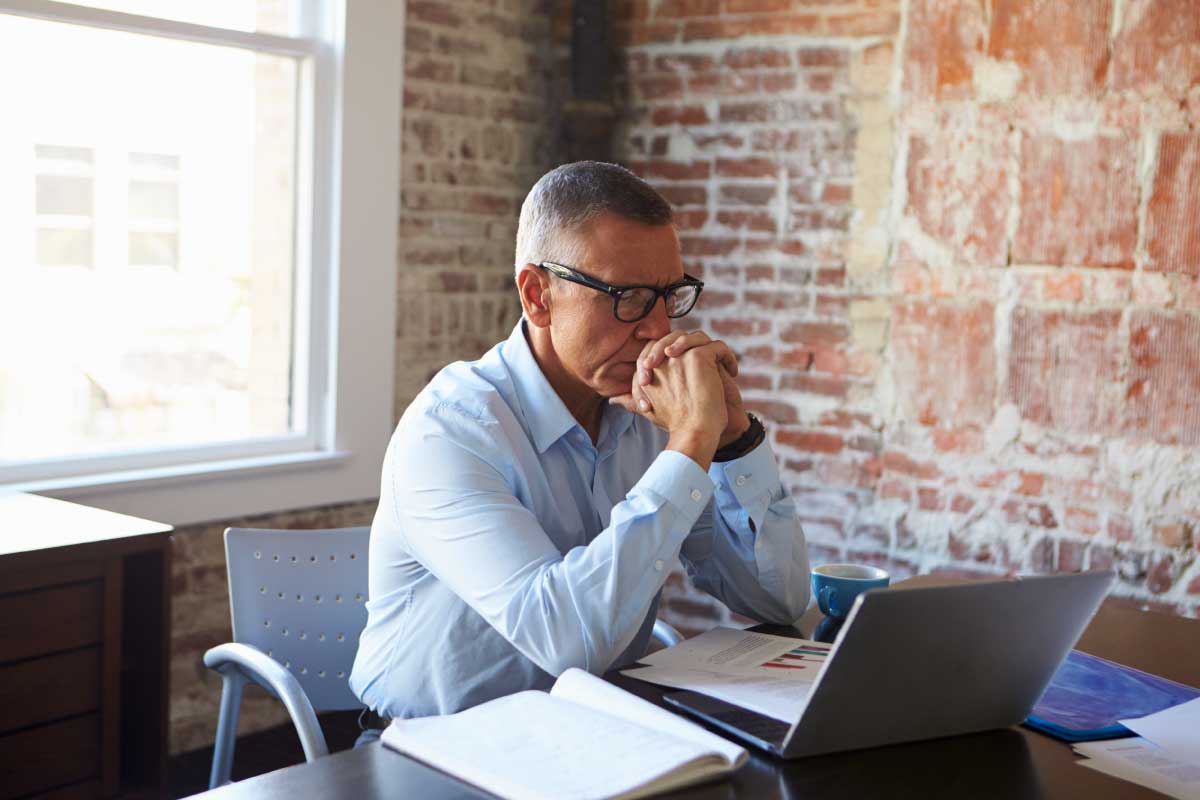 Hombre de negocios sentado al frente de un computador portatil, con las manos sobre su boca, analizando información.