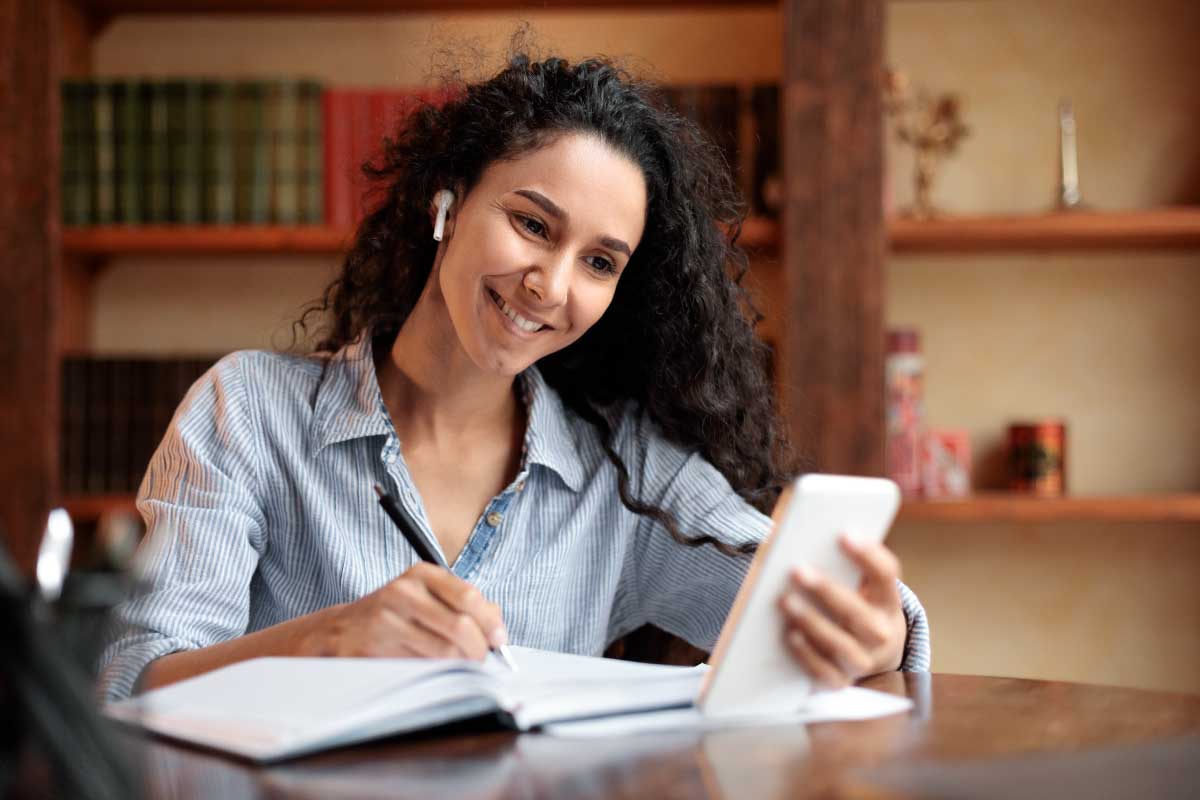 Mujer joven sonriendo, sentada en una mesa, escribiendo en una libreta y sosteniendo una tableta con la otra mano.