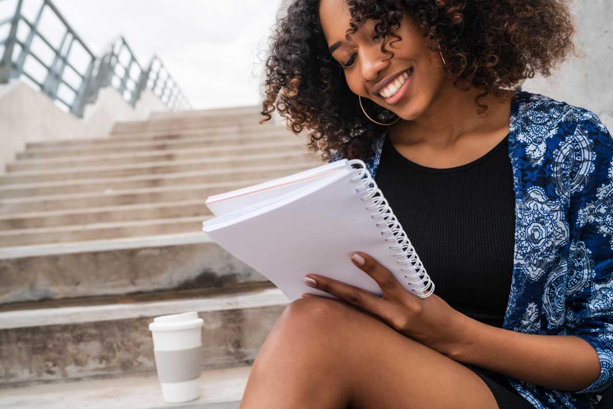 Mujer joven sonriente, sentada en una escalas tomando notas en un cuaderno.