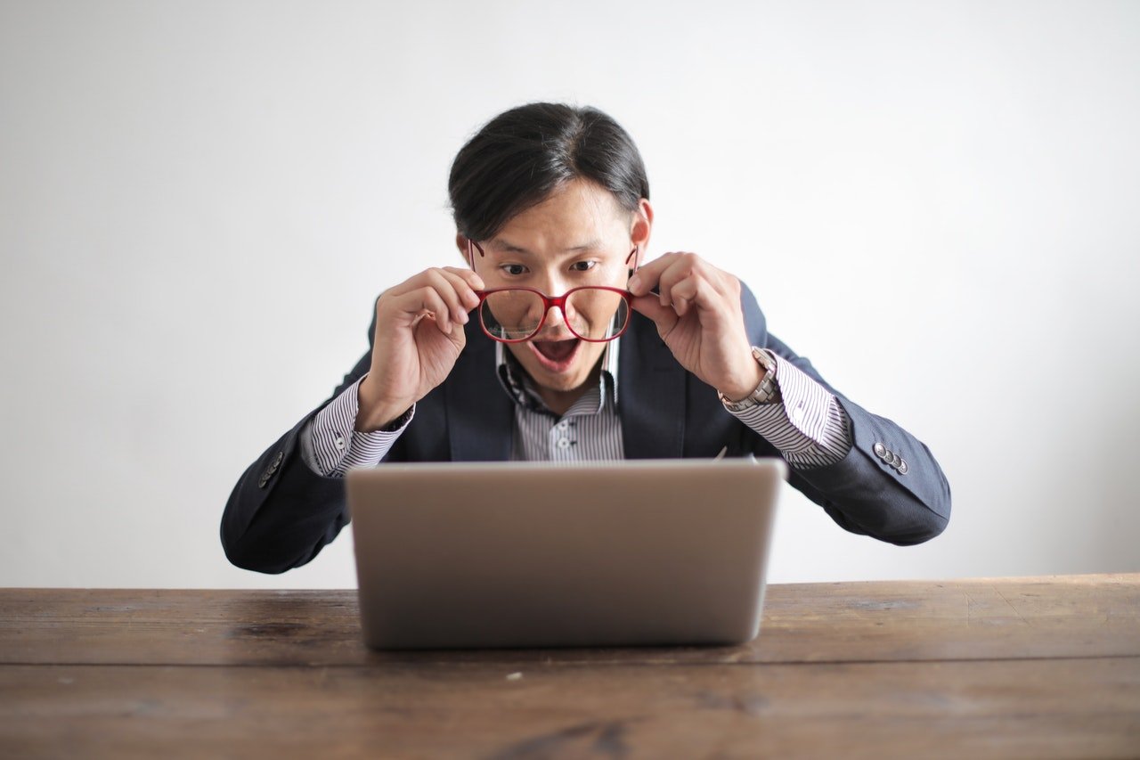 Hombre sentado al frente de un computador portatil removiendo sus lentes y con expresión de sorpresa.