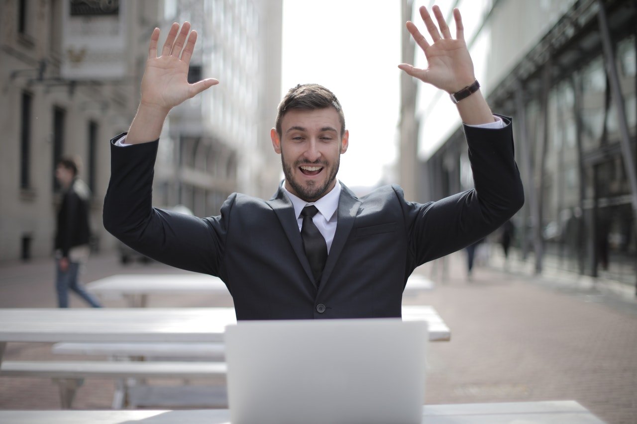 Hombre de negocios sentado en el exterior al frente de un computador con los brazos levantados en señal de felicidad